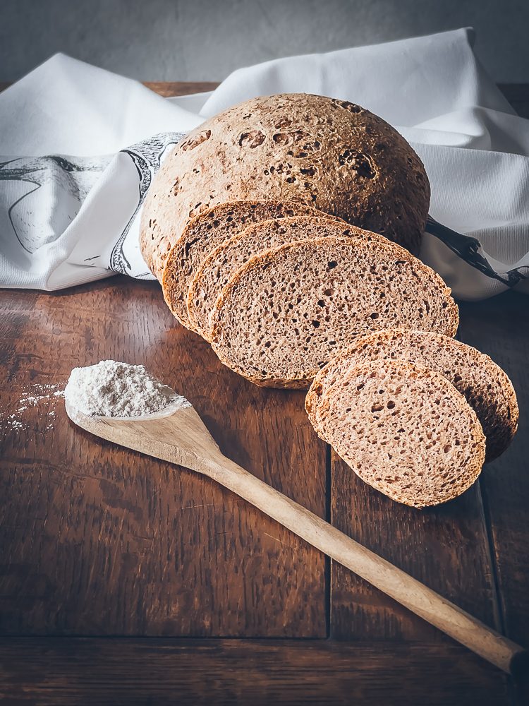 Auf einem Holztisch liegen aufgeschnittene Brotscheiben, außerdem eine  weiße Serviette und ein Holzlöffel gefüllt mit Mehl.